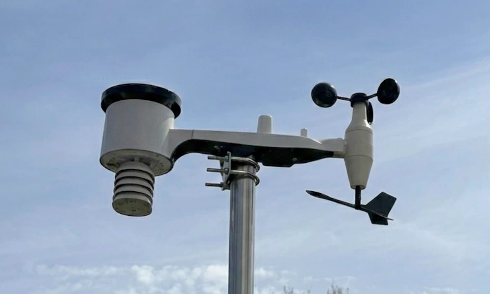 Eine Nahaufnahme einer an einem Metallmast montierten integrierten Wetterstation vor klarem blauen Himmel. Die weiße Einheit hat auf der linken Seite einen Regenmesser mit darunter liegenden Belüftungslamellen und auf der rechten Seite ein Anemometer (Schalenrad) und eine Windfahne